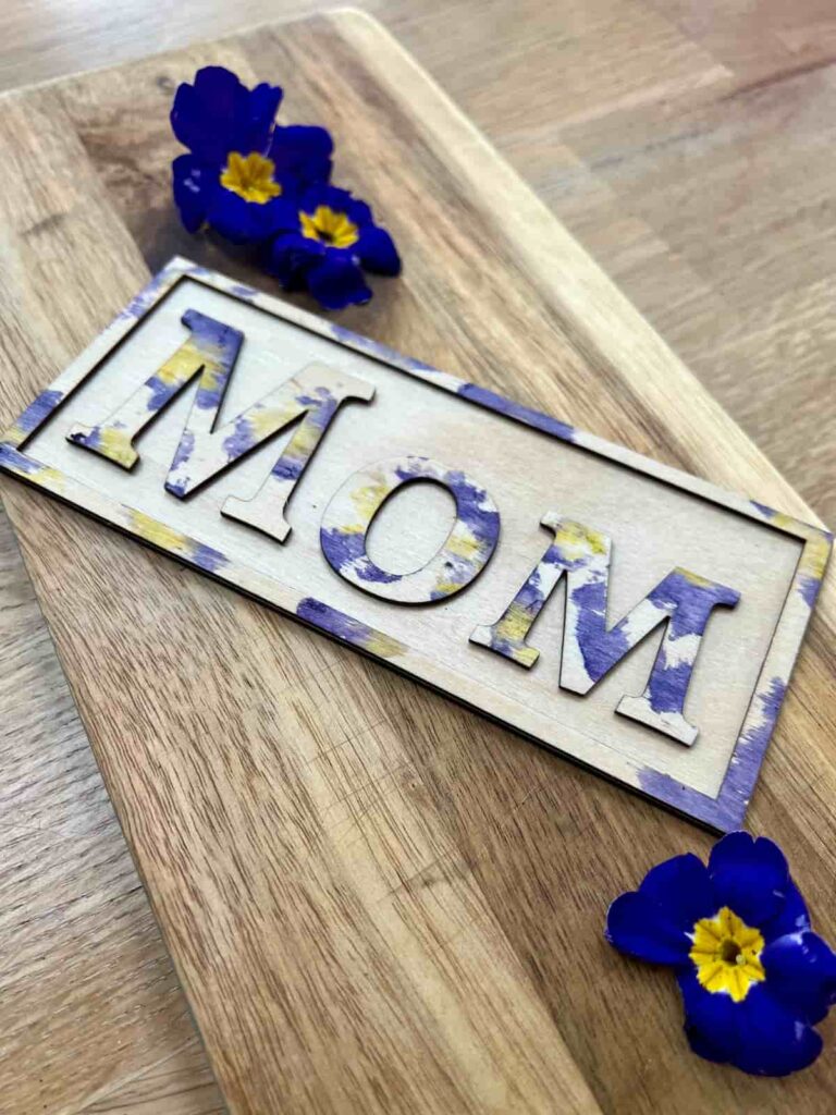 image shows wooden letters of the word mom with purple flowers.