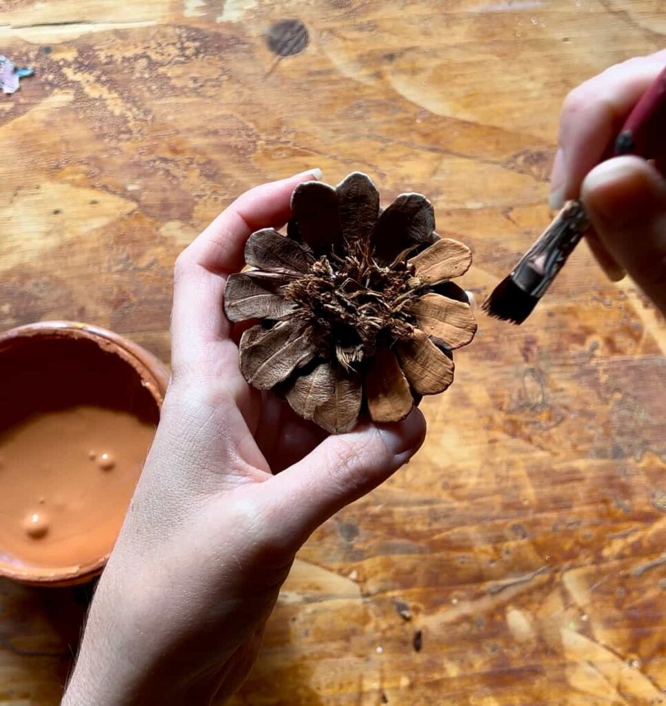 image shows hand painting a pinecone with orange paint.