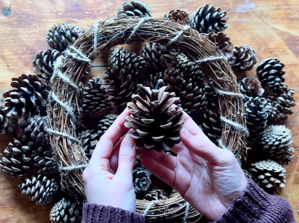 image shows hands holding a pinecone with wreath below.