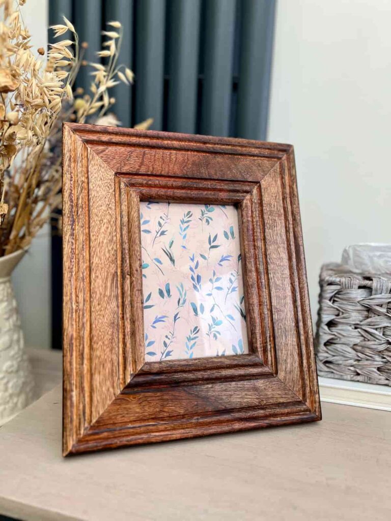 image shows wooden photo frame on a table next to flowers.